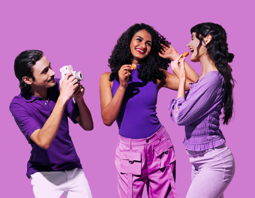 Three young adults enjoying snacks against a purple background; one man taking a photo while two women smile and hold food.