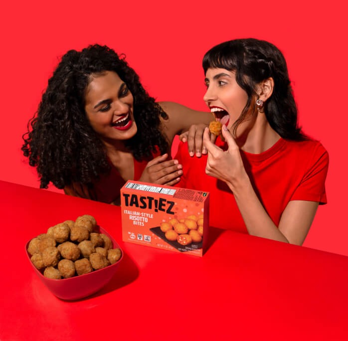 Two women laughing and enjoying tasting Italian-style risotto bites from a bowl and box labeled TASTIEZ on a red table and background.